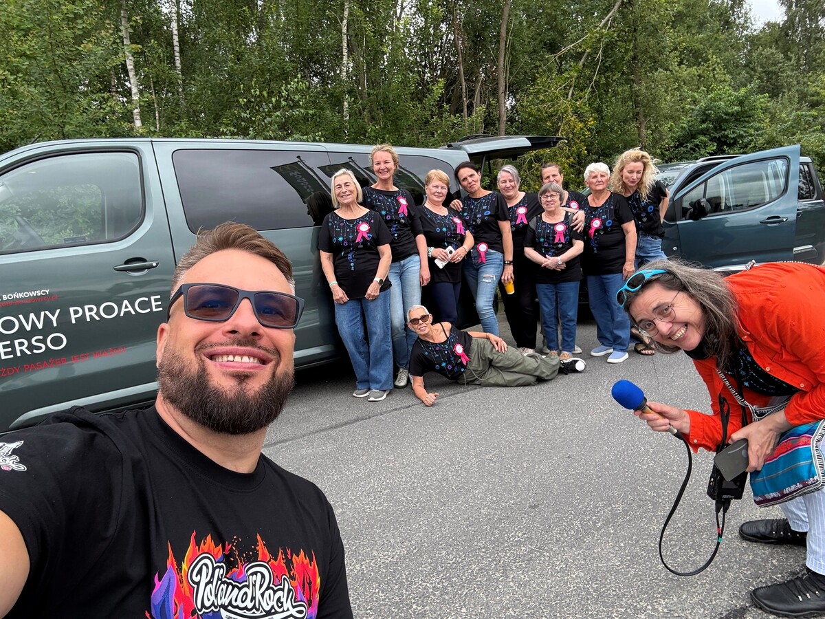 A group of women pose for a group photo near a parked Toyota Proace bus. In the foreground, a man wearing a Pol'and'Rock T-shirt takes a selfie, while a woman with a microphone leans into the frame.